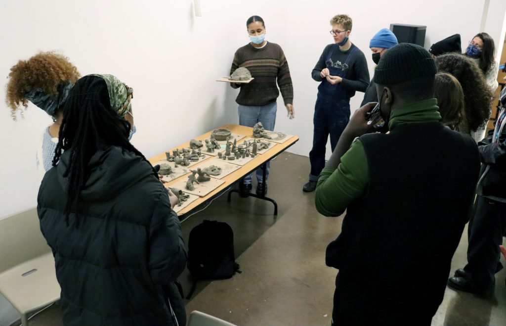Photo of 10 people, including the Art Assassins and session facilitators, standing around a table of clay sculptures. They are in discussion. Many are wearing face masks. 